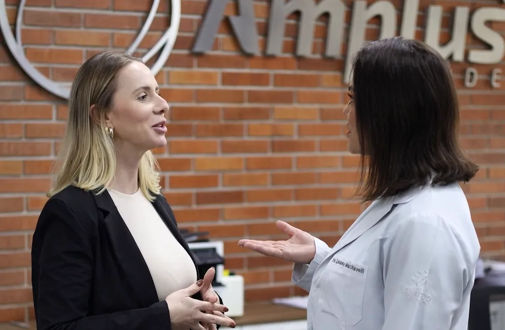 Duas profissionais da Amplus conversando na recepção da clínica, em frente à parede com a marca
